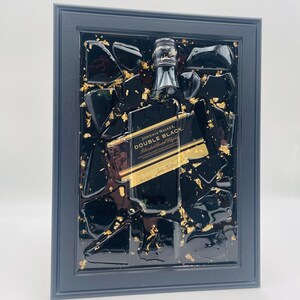 以下が含まれることがあります： ジョニーウォーカーダブルブラックのボトルをあしらった額装アート。ボトルは黒い破片と金箔のアクセントに囲まれ、黒いフレームに収められています。ラベルが見え、アートワークはモダンな美学を持っています。