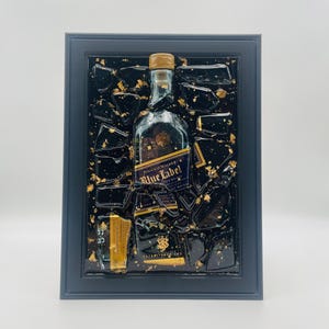 May include: A framed artwork featuring a Johnnie Walker Blue Label bottle. The bottle is set against a black background with gold leaf accents. The frame is a dark gray color, creating a striking contrast.