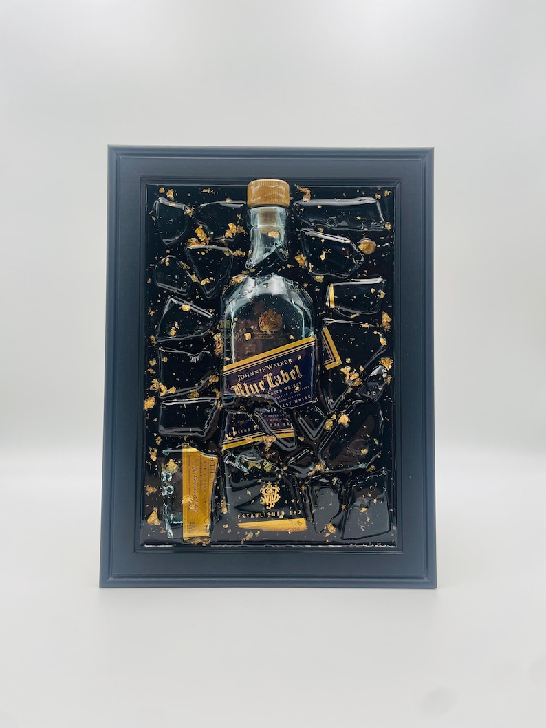 May include: A framed artwork featuring a Johnnie Walker Blue Label bottle. The bottle is set against a black background with gold leaf accents. The frame is a dark gray color, creating a striking contrast.