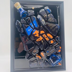May include: A framed artwork featuring fragments of a blue bottle with gold accents and orange highlights. The artwork is set against a black background within a gray frame. The bottle fragments are arranged in a scattered pattern.