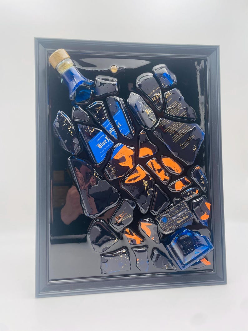 May include: A framed artwork featuring fragments of a blue bottle with gold accents and orange highlights. The artwork is set against a black background within a gray frame. The bottle fragments are arranged in a scattered pattern.