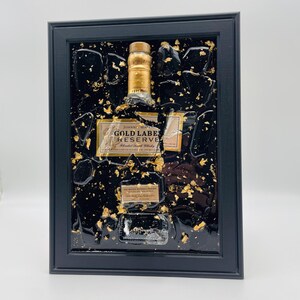 May include: Framed artwork featuring a Johnnie Walker Gold Label Reserve bottle. The bottle is set against a black background with gold leaf accents. The frame is black and rectangular, creating a striking contrast.