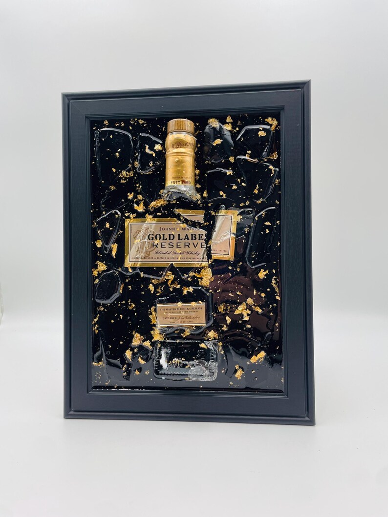 May include: Framed artwork featuring a Johnnie Walker Gold Label Reserve bottle. The bottle is set against a black background with gold leaf accents. The frame is black and rectangular, creating a striking contrast.