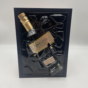 Pu&ograve; includere: Un'opera d'arte incorniciata con una bottiglia e un'etichetta di whisky Johnnie Walker Gold Label Reserve, su sfondo nero. Il tappo dorato e l'etichetta della bottiglia sono visibili, all'interno di una cornice nera.