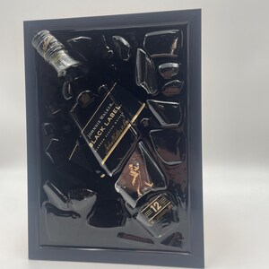 May include: A framed artwork featuring a Johnnie Walker Black Label bottle and fragments. The bottle is black with gold lettering and a label that reads "12 Years". The artwork is set against a black background within a dark blue frame.