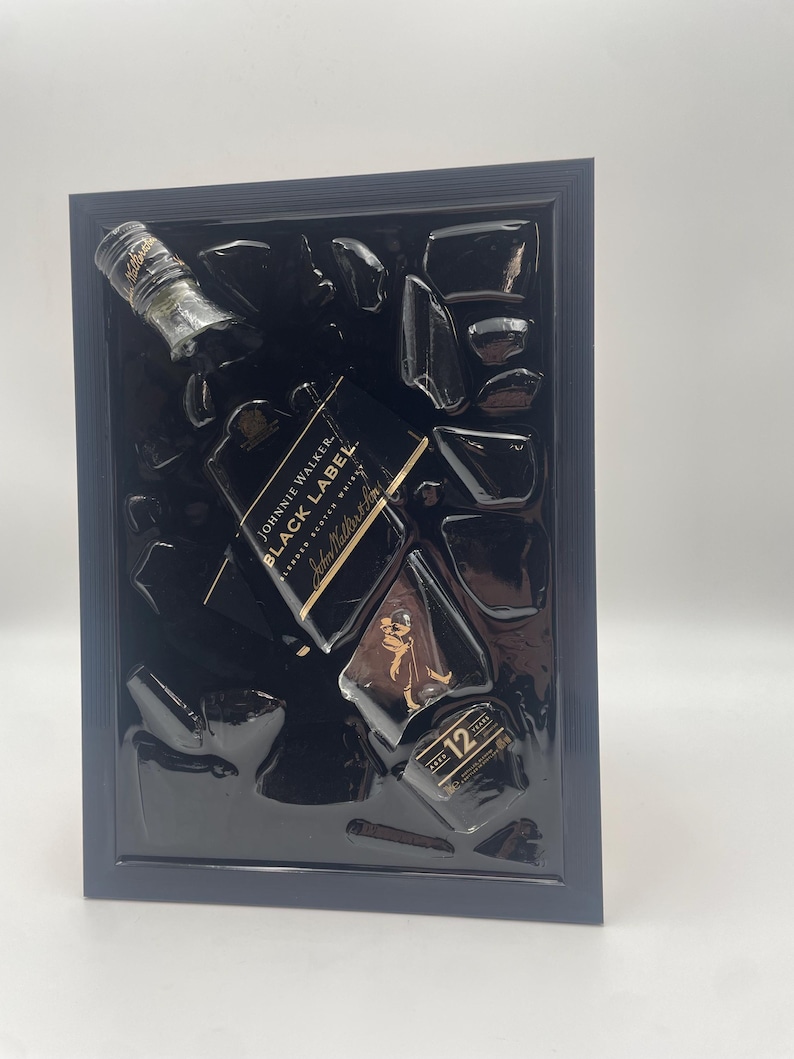 May include: A framed artwork featuring a Johnnie Walker Black Label bottle and fragments. The bottle is black with gold lettering and a label that reads "12 Years". The artwork is set against a black background within a dark blue frame.