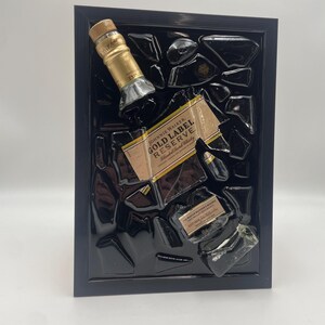 Pu&ograve; includere: Un'opera d'arte incorniciata con una bottiglia di Johnnie Walker Gold Label Reserve parzialmente rotta. L'etichetta e il tappo sono visibili, su sfondo nero all'interno di una cornice nera. L'opera ha un'estetica moderna unica.