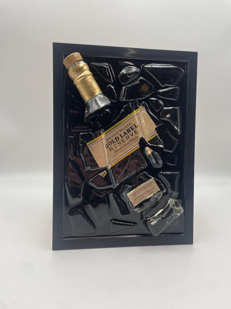 Pu&ograve; includere: Un'opera d'arte incorniciata con una bottiglia di Johnnie Walker Gold Label Reserve parzialmente rotta. L'etichetta e il tappo sono visibili, su sfondo nero all'interno di una cornice nera. L'opera ha un'estetica moderna unica.