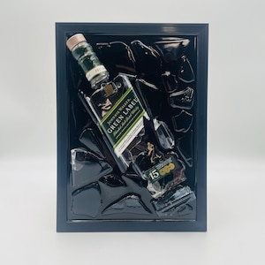 May include: A framed artwork featuring a Johnnie Walker Green Label whisky bottle, with a 15-year label, and fragments of what appears to be broken glass. The artwork is set against a black background within a black frame.