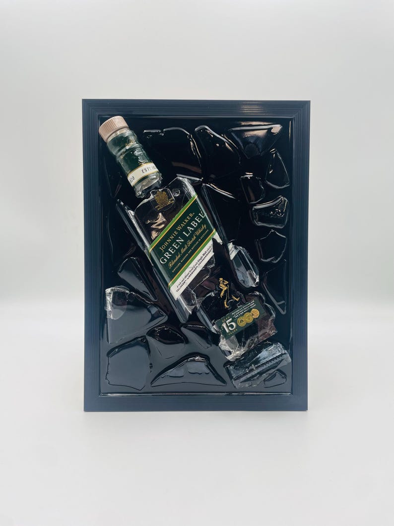 May include: A framed artwork featuring a Johnnie Walker Green Label whisky bottle, with a 15-year label, and fragments of what appears to be broken glass. The artwork is set against a black background within a black frame.