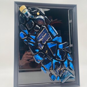 Op de afbeelding: Een ingelijst kunstwerk met fragmenten van een Johnnie Walker Blue Label fles. De stukken zijn in een verspreid patroon gerangschikt, met blauwe en zwarte tinten. De lijst is donkergrijs.