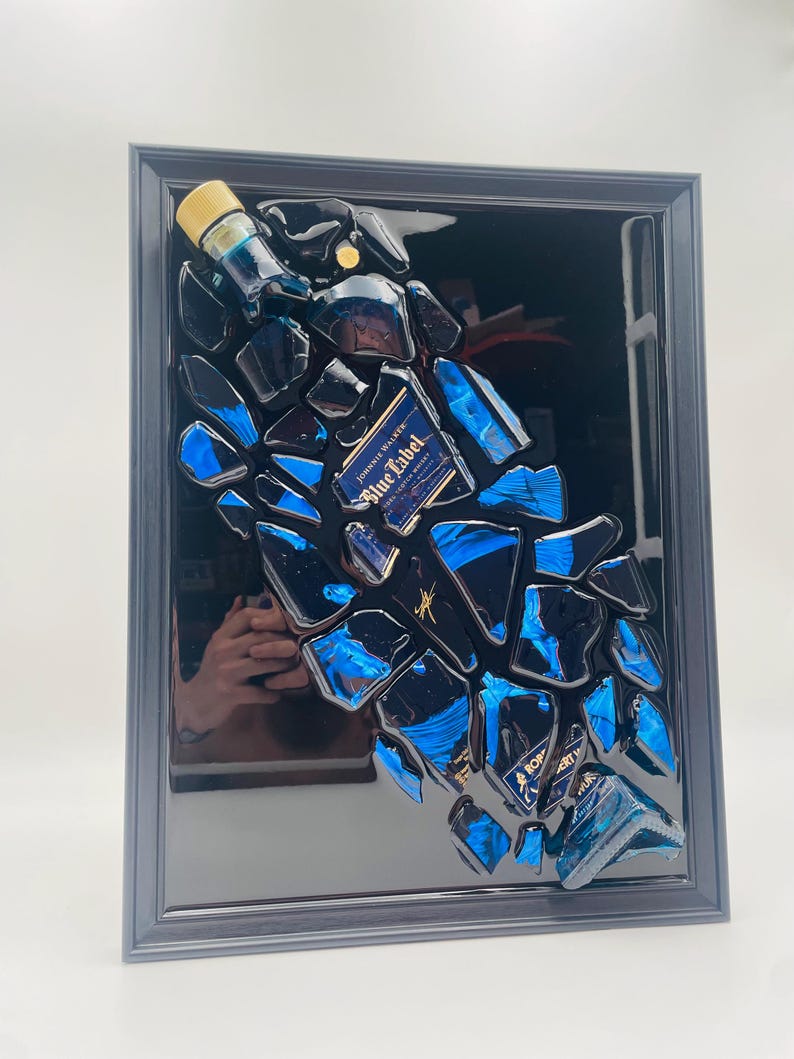 Op de afbeelding: Een ingelijst kunstwerk met fragmenten van een Johnnie Walker Blue Label fles. De stukken zijn in een verspreid patroon gerangschikt, met blauwe en zwarte tinten. De lijst is donkergrijs.