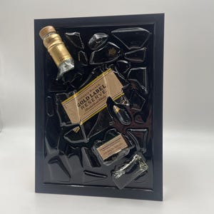 Pu&ograve; includere: Un'opera d'arte incorniciata con frammenti di una bottiglia di Johnnie Walker Gold Label Reserve. L'opera include il tappo e l'etichetta della bottiglia, su uno sfondo nero all'interno di una cornice nera. L'etichetta recita "Gold Label Reserve".