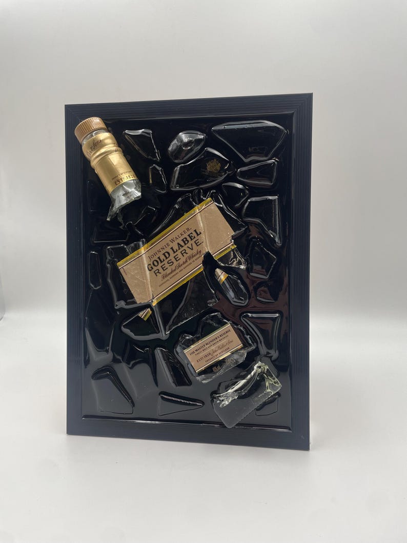 Pu&ograve; includere: Un'opera d'arte incorniciata con frammenti di una bottiglia di Johnnie Walker Gold Label Reserve. L'opera include il tappo e l'etichetta della bottiglia, su uno sfondo nero all'interno di una cornice nera. L'etichetta recita "Gold Label Reserve".