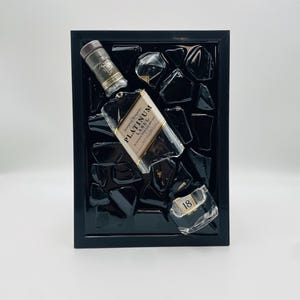 Op de afbeelding: Een ingelijst kunstwerk met een Johnnie Walker Platinum Label whiskyfles, dop en een stuk van de fles met het nummer 18. De objecten zijn geplaatst tegen een achtergrond van zwarte glasstukken in een zwart frame.