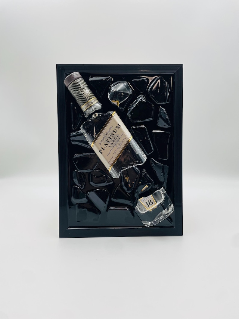 Op de afbeelding: Een ingelijst kunstwerk met een Johnnie Walker Platinum Label whiskyfles, dop en een stuk van de fles met het nummer 18. De objecten zijn geplaatst tegen een achtergrond van zwarte glasstukken in een zwart frame.