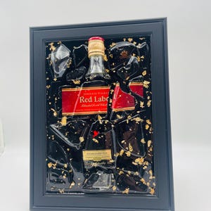 K&ouml;nnte beinhalten: Gerahmtes Kunstwerk mit einer Johnnie Walker Red Label Flasche, mit rotem Etikett und goldenen Akzenten. Die Flasche ist in schwarzem Harz mit Blattgold eingeschlossen, eingebettet in einen schwarzen Rahmen. Der Text "ESTABLISHED 1820" ist sichtbar.
