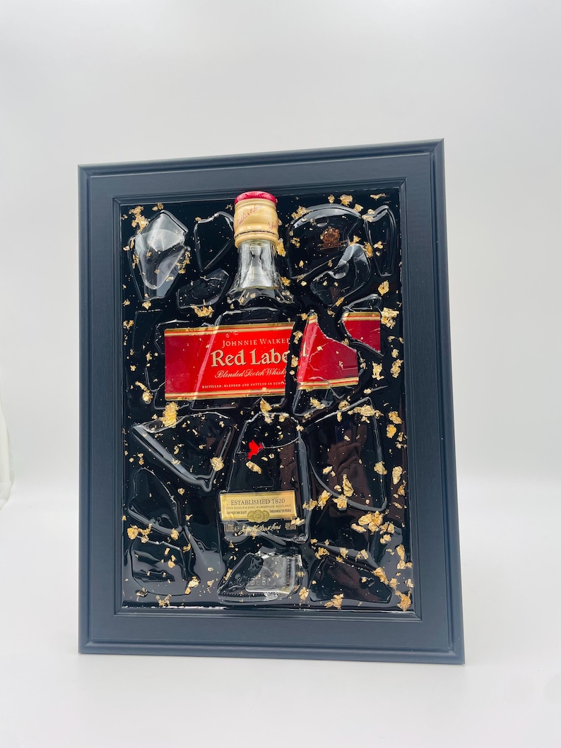 K&ouml;nnte beinhalten: Gerahmtes Kunstwerk mit einer Johnnie Walker Red Label Flasche, mit rotem Etikett und goldenen Akzenten. Die Flasche ist in schwarzem Harz mit Blattgold eingeschlossen, eingebettet in einen schwarzen Rahmen. Der Text "ESTABLISHED 1820" ist sichtbar.