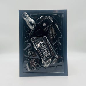 Pode incluir: Uma obra de arte emoldurada com uma coleção de garrafas e fragmentos de uísque Jack Daniel's. As garrafas são pretas e transparentes, com rótulos brancos. A obra de arte está em um fundo preto dentro de uma moldura azul escura.