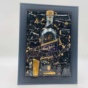 May include: Framed artwork featuring a Johnnie Walker Blue Label bottle and fragments, set against a black background with gold leaf accents. The artwork is encased in a dark blue frame, creating a striking visual contrast.