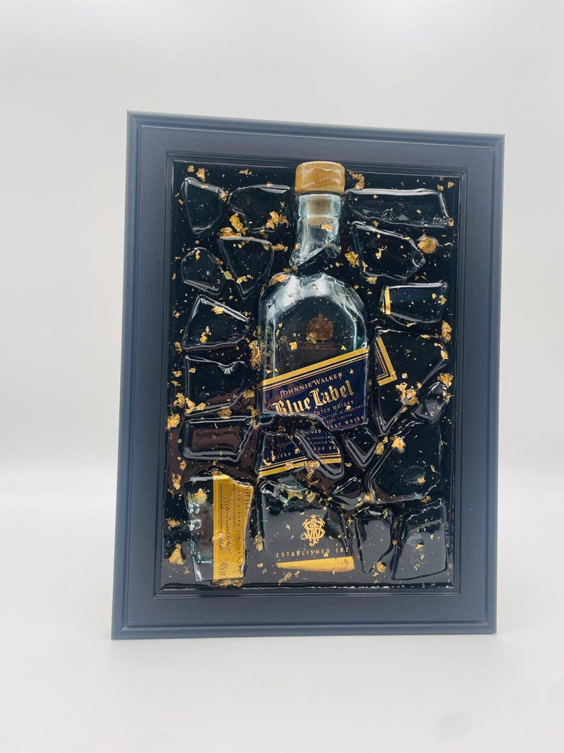 May include: Framed artwork featuring a Johnnie Walker Blue Label bottle and fragments, set against a black background with gold leaf accents. The artwork is encased in a dark blue frame, creating a striking visual contrast.