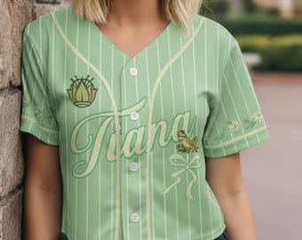 Op de afbeelding: Een lichtgroen honkbalshirt met verticale witte strepen. Het shirt heeft de naam "Tiana" in een scriptlettertype, samen met een kikker- en bloemenontwerp. Het shirt heeft een V-hals en een knoopsluiting.