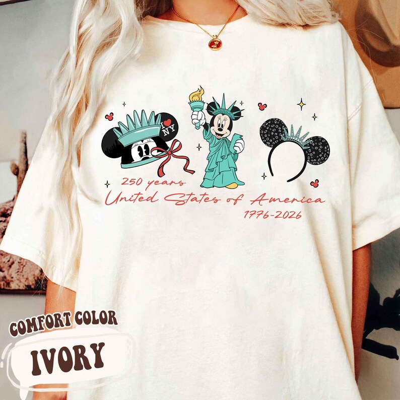 May include: Ivory Comfort Color t-shirt featuring Mickey Mouse in Statue of Liberty attire. The shirt has the text "250 years United States of America 1776-2026" in red script. The design includes Mickey Mouse ears with a crown.