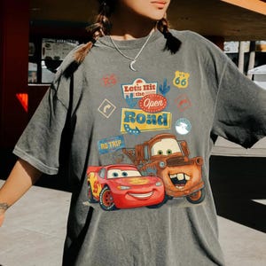 May include: Grey t-shirt featuring Lightning McQueen and Mater from the film Cars. The shirt has the text "Let's Hit the Open Road" with travel-themed graphics. The design is printed on a grey, oversized tee.