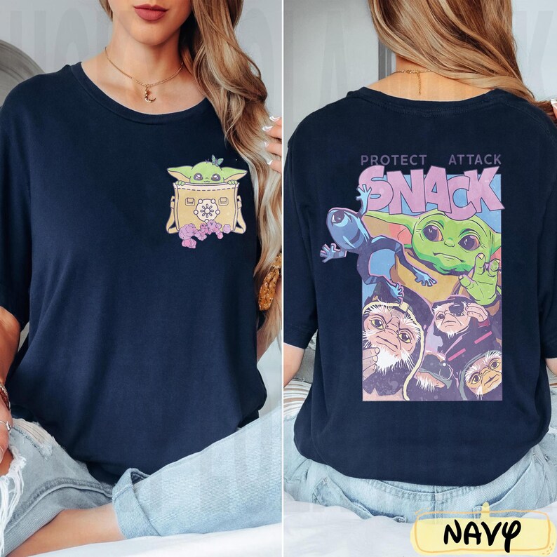 May include: Navy blue t-shirt featuring a graphic of a green alien character in a yellow carrier on the front. The back of the shirt has a colorful graphic with the text "PROTECT ATTACK SNACK" and other characters. The word "NAVY" is at the bottom.