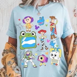 May include: Light blue t-shirt featuring colorful cartoon character illustrations. The design includes characters like Woody, Buzz Lightyear, and Jessie, with the names of the characters written in cursive. The shirt has a crew neck and short sleeves.