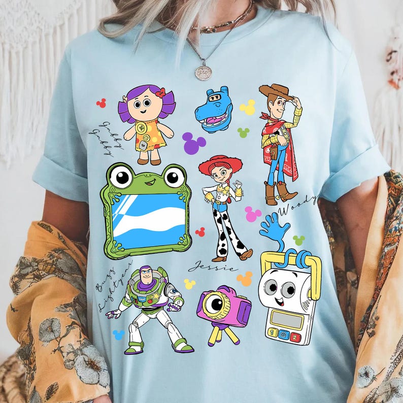 May include: Light blue t-shirt featuring colorful cartoon character illustrations. The design includes characters like Woody, Buzz Lightyear, and Jessie, with the names of the characters written in cursive. The shirt has a crew neck and short sleeves.