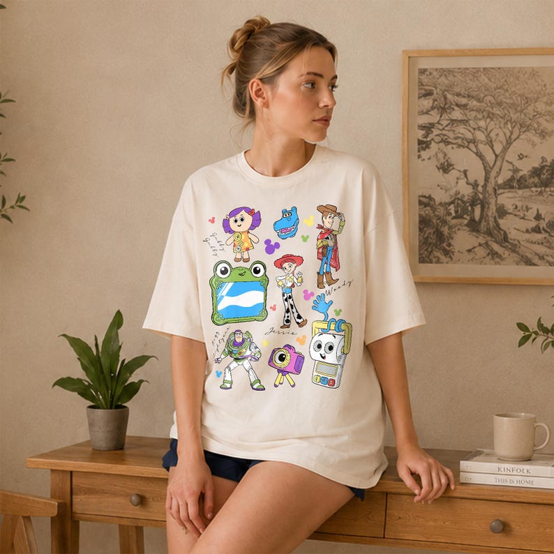 May include: Cream-colored t-shirt featuring colorful cartoon character illustrations. The shirt has short sleeves and a crew neck. The graphics include a doll, a blue dinosaur, a cowboy, a frog, a cowgirl, a space ranger, and a camera.