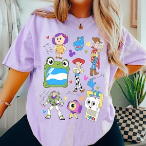 May include: A light purple t-shirt featuring colorful cartoon character illustrations. The shirt showcases characters like Woody, Buzz Lightyear, and Jessie, with character names and decorative elements. The design is playful and vibrant.