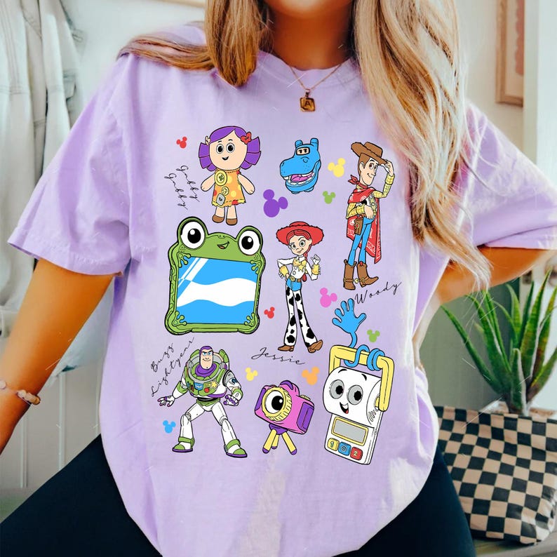 May include: A light purple t-shirt featuring colorful cartoon character illustrations. The shirt showcases characters like Woody, Buzz Lightyear, and Jessie, with character names and decorative elements. The design is playful and vibrant.