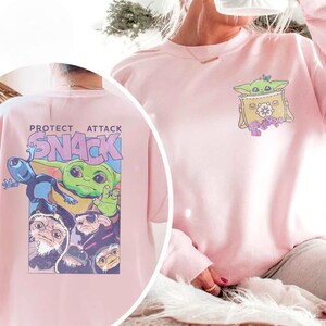 May include: Pink sweatshirt featuring a graphic of a green alien character in a yellow bag on the front. The back of the sweatshirt has the text "PROTECT ATTACK SNACK" with a graphic of the same alien character and other characters.