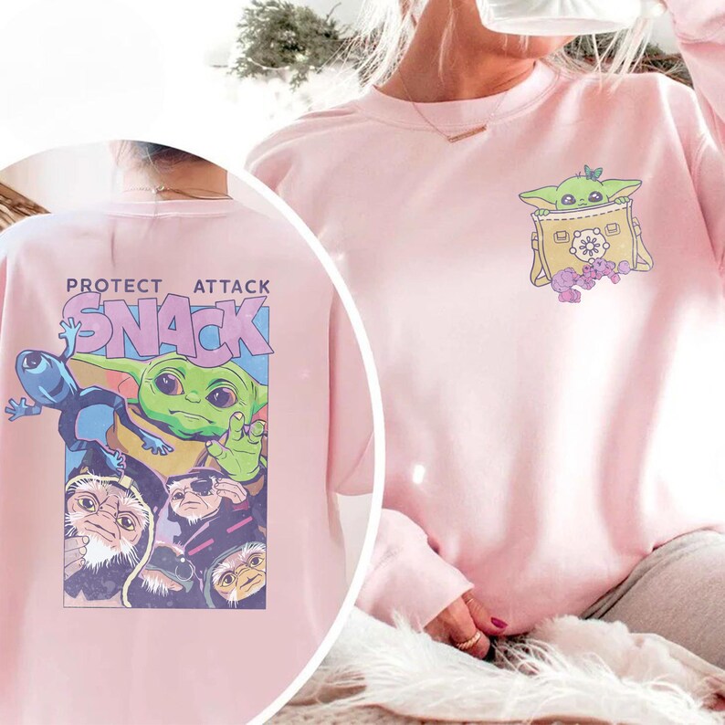 May include: Pink sweatshirt featuring a graphic of a green alien character in a yellow bag on the front. The back of the sweatshirt has the text "PROTECT ATTACK SNACK" with a graphic of the same alien character and other characters.