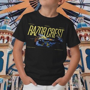 May include: Black t-shirt featuring the text "RAZOR CREST" in yellow, with a blue and yellow spaceship graphic. The shirt also includes text that reads "HYPERSPACE CAPABILITIES" and "BOUNTY HUNTER TRANSPORT".