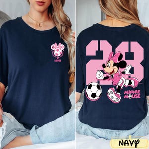 May include: Navy blue t-shirt with a pink Minnie Mouse soccer design. The front has a soccer ball with the number 28 and "Minnie". The back shows Minnie Mouse in a soccer uniform with the number 28 and a soccer ball.