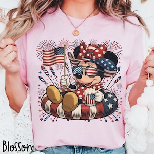 May include: Pink t-shirt featuring a patriotic Minnie Mouse design. Minnie is wearing sunglasses, a bow, and holding an American flag and a mug. The design includes fireworks and a red, white, and blue inner tube. The word "Blossom" is at the bottom.