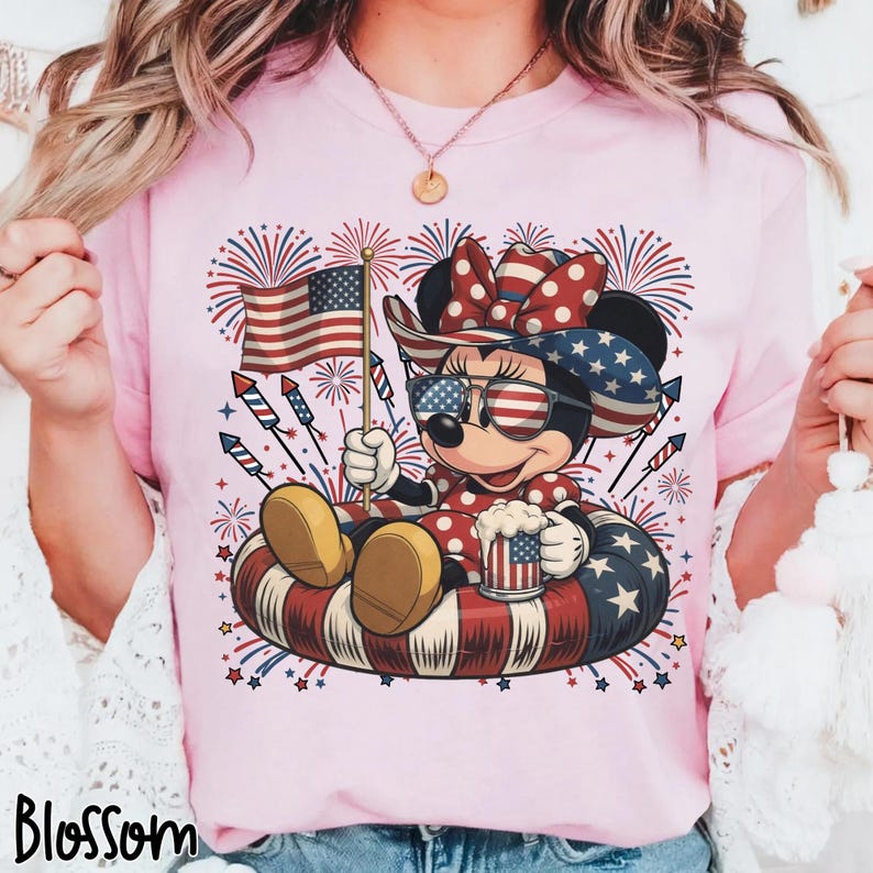 May include: Pink t-shirt featuring a patriotic Minnie Mouse design. Minnie is wearing sunglasses, a bow, and holding an American flag and a mug. The design includes fireworks and a red, white, and blue inner tube. The word "Blossom" is at the bottom.