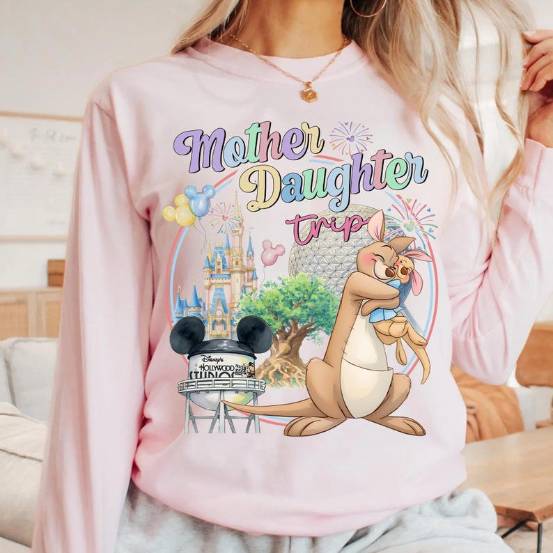 May include: Pale pink long-sleeved top featuring a colourful graphic. The design includes the text "Mother Daughter Trip" with Disney illustrations of a kangaroo and joey, a castle, and a water tower.
