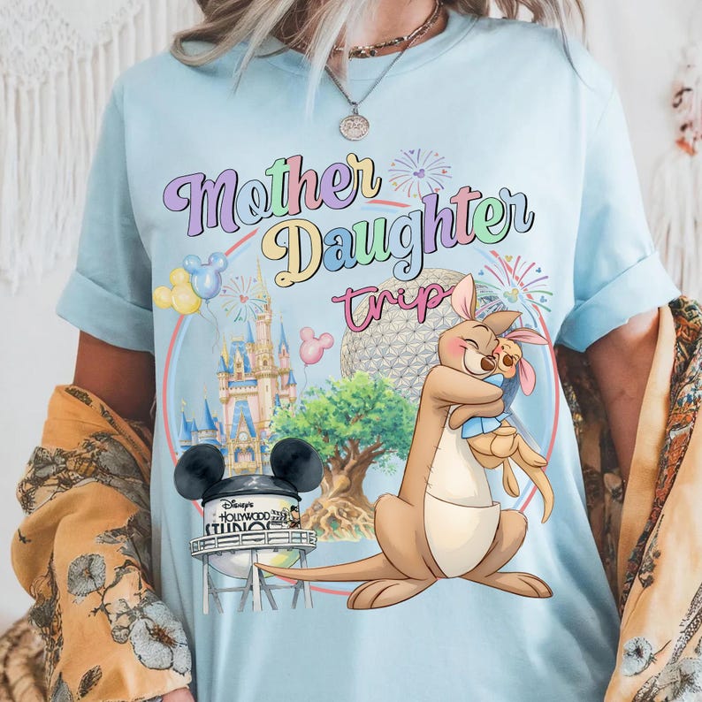 May include: Pale blue t-shirt with a colourful graphic that reads "Mother Daughter Trip". The design includes a kangaroo hugging a joey, a castle, and a water tower with Mickey Mouse ears. The shirt has rolled-up sleeves.