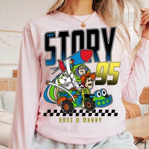May include: A light pink long-sleeve sweatshirt featuring a graphic from the movie Toy Story. The graphic includes Buzz Lightyear and Woody in a race car with the text "TOY STORY 95" and "BUZZ & WOODY".