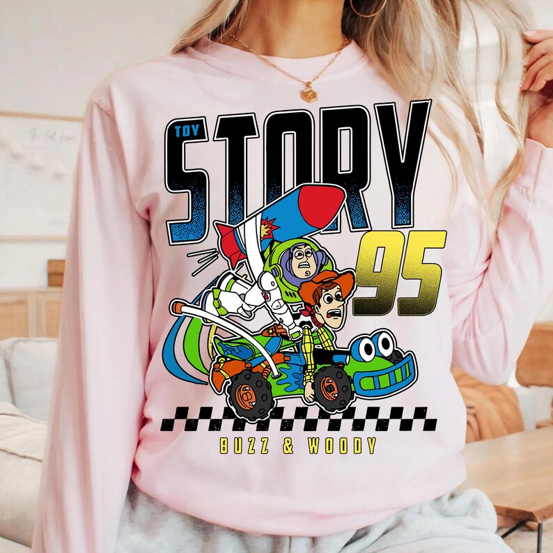 May include: A light pink long-sleeve sweatshirt featuring a graphic from the movie Toy Story. The graphic includes Buzz Lightyear and Woody in a race car with the text "TOY STORY 95" and "BUZZ & WOODY".
