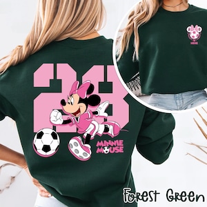 May include: Forest green sweatshirt with a pink graphic of Minnie Mouse playing football, with the number 23 in pink. The text "MINNIE MOUSE" is below the graphic. A smaller graphic of a football and the text "2028 Minnie" is on the upper right.