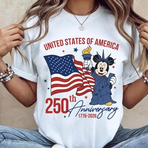 May include: White t-shirt featuring a graphic celebrating the 250th anniversary of the United States of America. The design includes the American flag, a cartoon character dressed as the Statue of Liberty, and the text "United States of America" and "250th Anniversary 1776-2026".