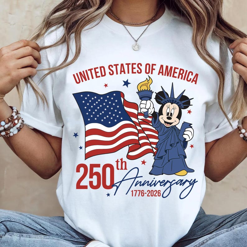 May include: White t-shirt featuring a graphic celebrating the 250th anniversary of the United States of America. The design includes the American flag, a cartoon character dressed as the Statue of Liberty, and the text "United States of America" and "250th Anniversary 1776-2026".