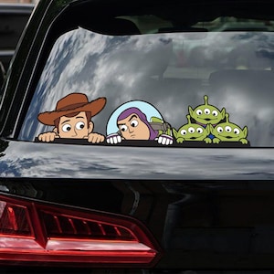 May include: A car window decal featuring characters from the animated film Toy Story. The decal shows Woody in a cowboy hat, Buzz Lightyear, and three green aliens peeking over a surface. The characters are in a cartoon style with bold outlines and bright colors.