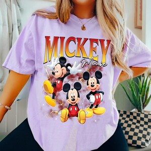 May include: A lilac t-shirt with three Mickey Mouse illustrations in red, yellow, and black. The text "Mickey Mouse" is printed in a gradient yellow and red font above the illustrations.