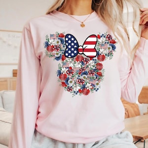 May include: A light pink long-sleeve sweatshirt featuring a floral design in the shape of Mickey Mouse ears. The design includes a bow with a stars and stripes pattern, and red, white, and blue flowers and American flags.
